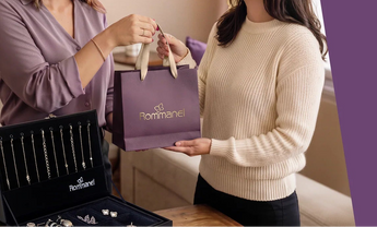 Person handing a purple Rommanel shopping bag to a customer next to a jewelry display case.