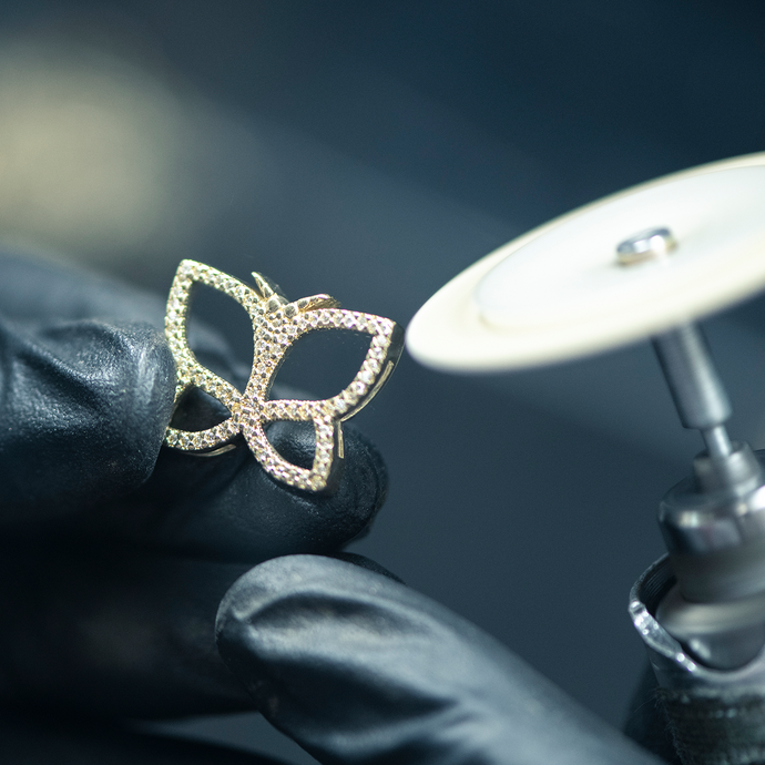 A jeweler wearing black gloves holds a gold pavé butterfly pendant next to a rotary polishing tool.