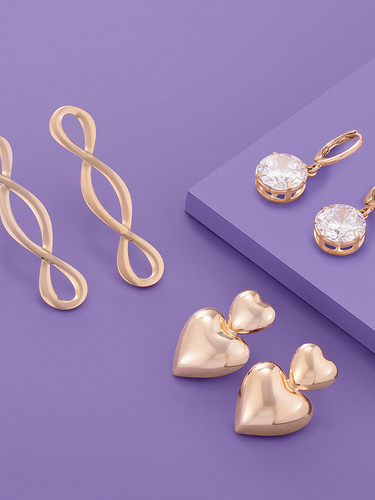 Product display banner on a light purple background showcasing three styles of gold earrings: a pair of wavy infinity-twist drop earrings, a pair of double-heart drop earrings, and a pair of gold huggie hoops featuring large round cubic zirconia stones.