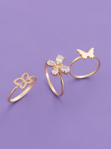 Vertical banner showcasing three gold rings on a purple background: a pave crystal butterfly band ring, a center ring with a flower motif made of marquise stones, and a minimalist gold band with a smooth butterfly accent.