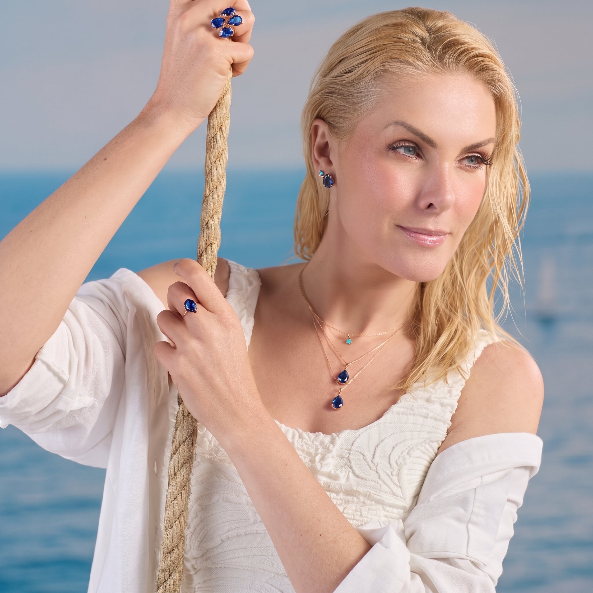 A blonde model poses against a serene ocean background while holding a rope, showcasing a gold fashion jewelry collection with deep blue teardrop gemstones—including layered necklaces, matching earrings, and a statement ring.