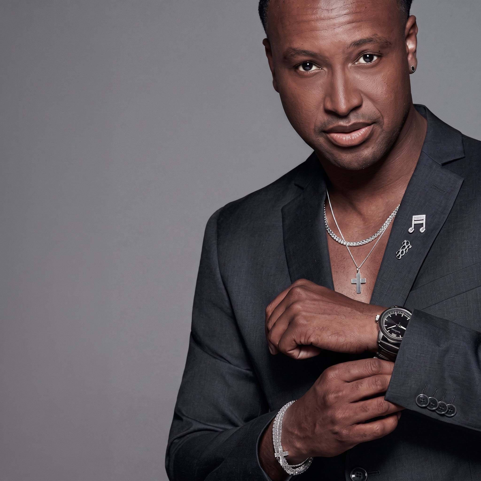 Half-body portrait of a man in a black blazer wearing sterling silver jewelry: a cross pendant necklace and stacked link bracelets. He holds his wrist, looking at the camera against a grey background.