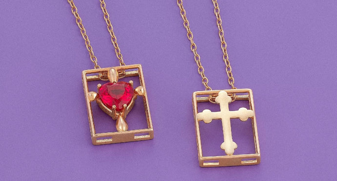 Close-up of two gold-toned pendant necklaces on a purple background. Each is a rectangular frame: the left pendant holds a large, prominent red heart-shaped gemstone, while the right pendant contains an empty, ornate cutout cross design.