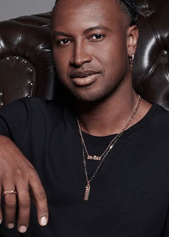 Man wearing layered gold necklaces with a "BEM-BLACK" pendant and gold vertical bar pendant, a cross hoop earring, and a gold ring.