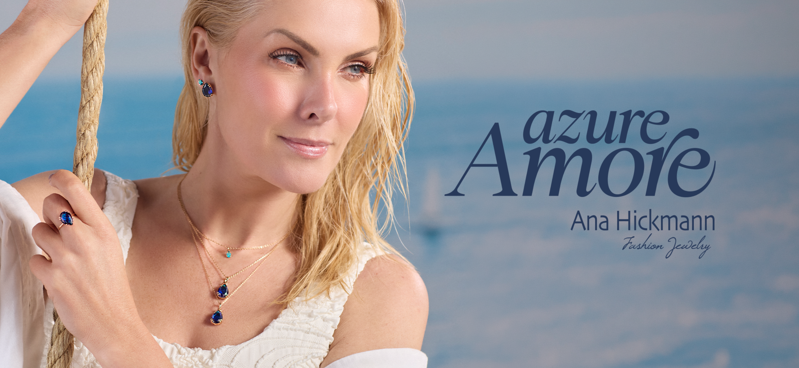 A blonde model poses against a serene ocean background while holding a rope, showcasing a gold fashion jewelry collection with deep blue teardrop gemstones—including layered necklaces, matching earrings, and a statement ring. Navy text on the right reads 