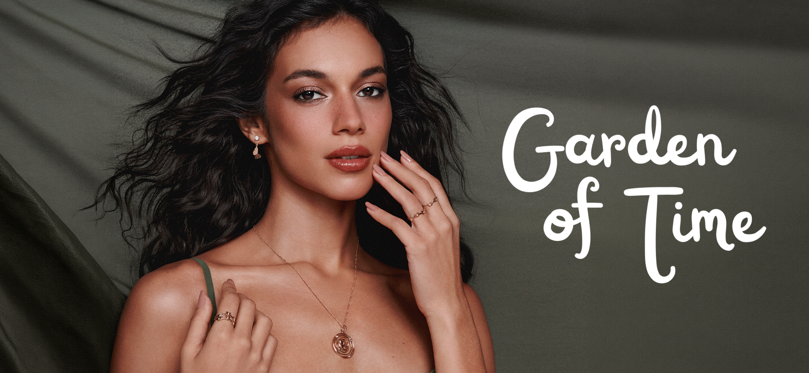 A model with dark, voluminous wavy hair and soft makeup poses against a muted olive-green backdrop, elegantly displaying delicate gold fine jewelry—including a pendant necklace, small drop earrings, and minimal stacked rings. White cursive script text on the right side of the image reads 