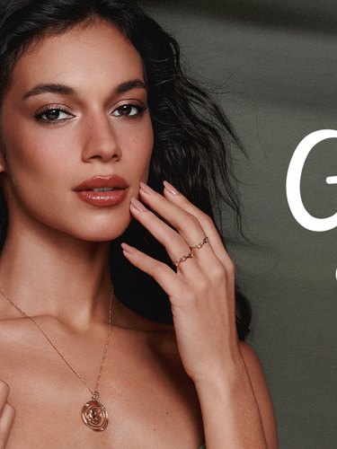 A model with dark, voluminous wavy hair and soft makeup poses against a muted olive-green backdrop, elegantly displaying delicate gold fine jewelry—including a pendant necklace, small drop earrings, and minimal stacked rings. White cursive script text on the right side of the image reads "Garden of Time."