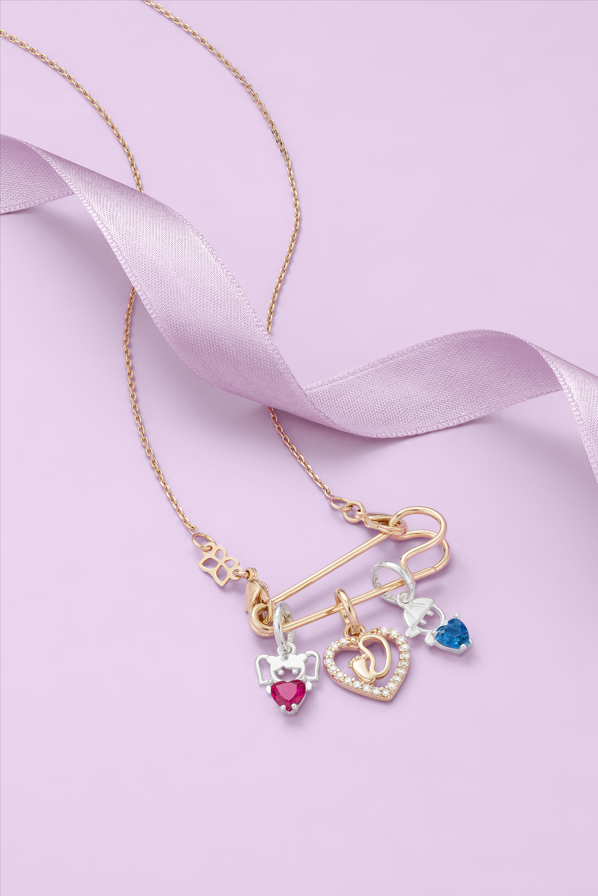 Gold safety pin necklace with various charms, including two heart-shaped ones, a blue one, and a baby's footprint charm on a light-purple background.